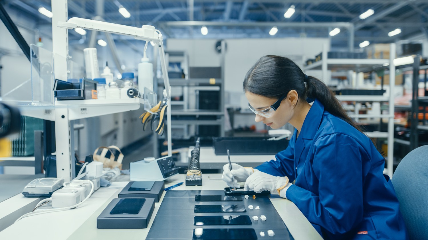 Young Female in Blue Work Coat is Assembling Printed Circuit Boards for Smartphones. Electronics Factory Workers in a High Tech Factory Facility. Photo: Shutterstock