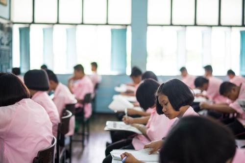 Male and female students dressed in pink uniforms sit at classroom desks taking exams.