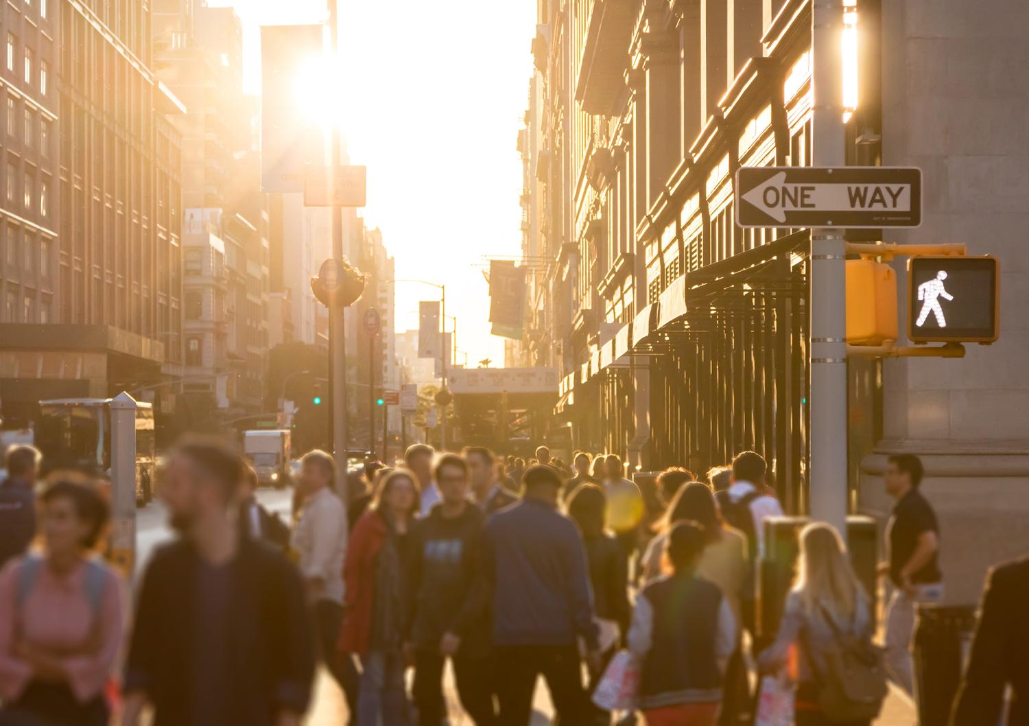 Diverse crowd of anonymous people walking down a busy street in Manhattan, New York City with bright sunlight shining in the background