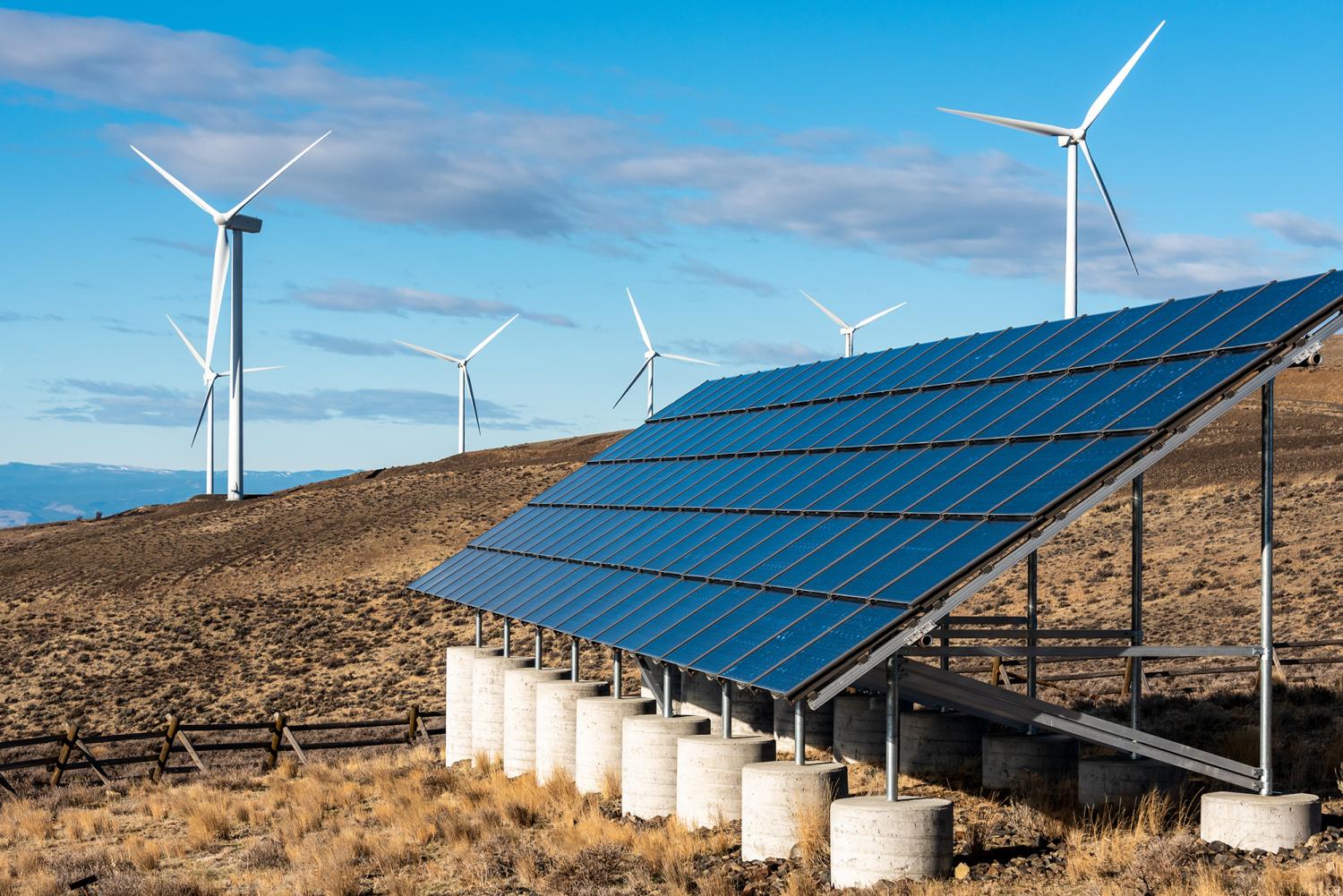 White wind turbines on a hill top, in a dry landscape with solar panels in foreground, blue sky and white clouds in background