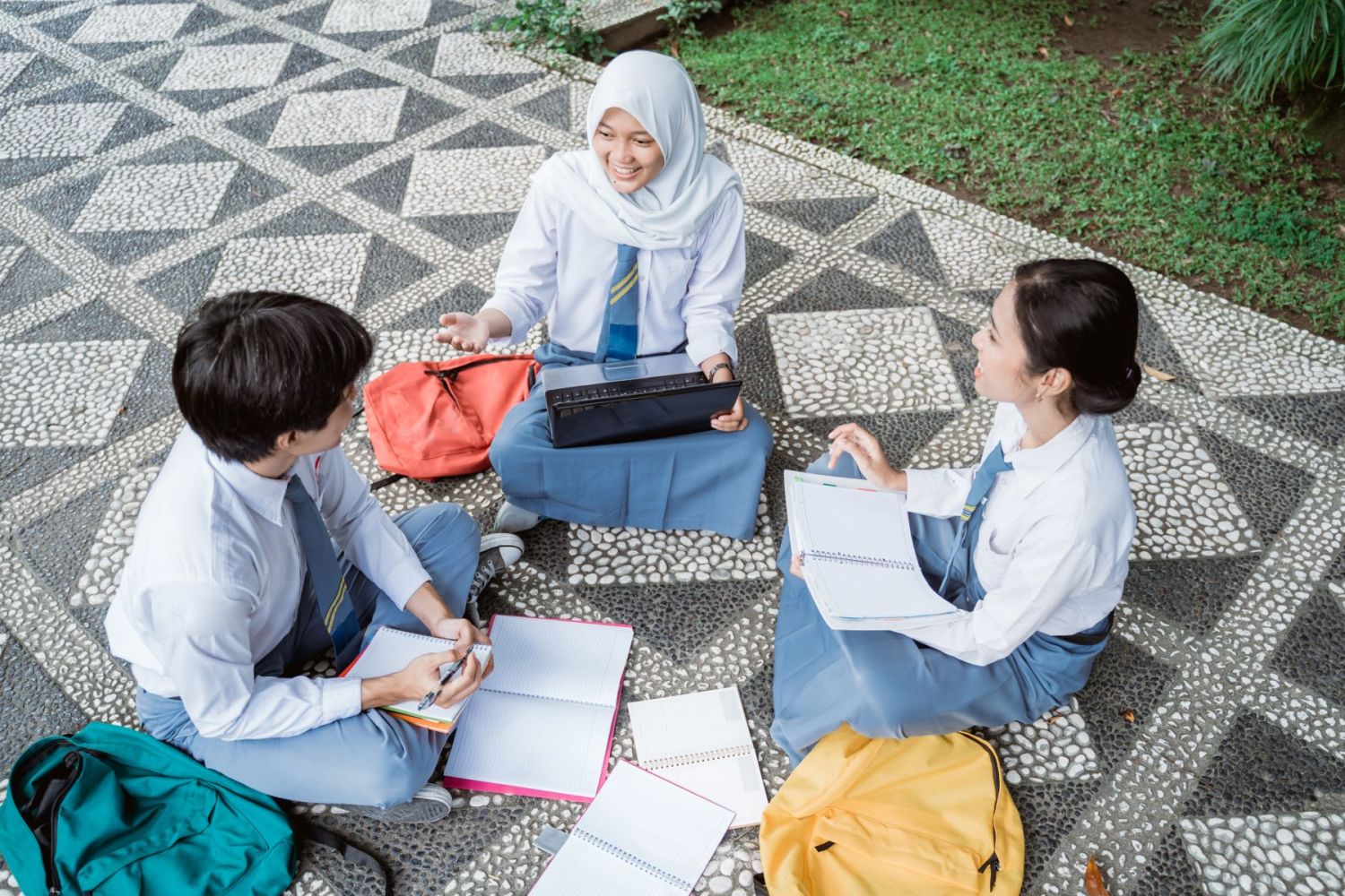 Three high school students sit on the ground outside studying and talking.