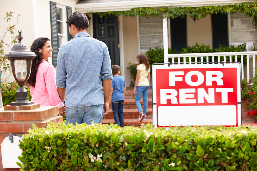 A family stands outside of a home with a "for rent" sign