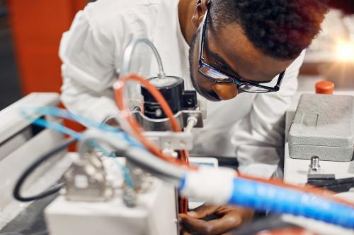 Portrait of Black man wearing white robe, working at LED factory, using tablet, connecting wires, programming equipment