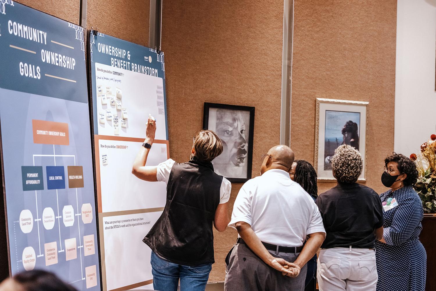 group of diverse individuals gather around poster board titled "Ownership & benefit Brainstorm." One woman holds marker while writing down ideas.