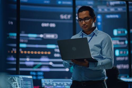 A young man works on a laptop computer in the middle of a room filled with computer monitors an controls