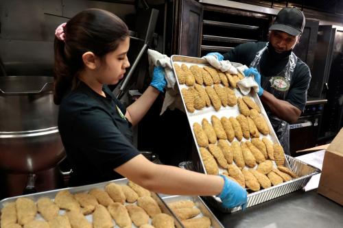 Prep workers Alejandra Vargas, left, and Tom Ross unload a tray of chicken nuggets from the oven, for asylum-seeking migrants at B.J’s Market and Bakery on Feb. 6, 2024, in Chicago.