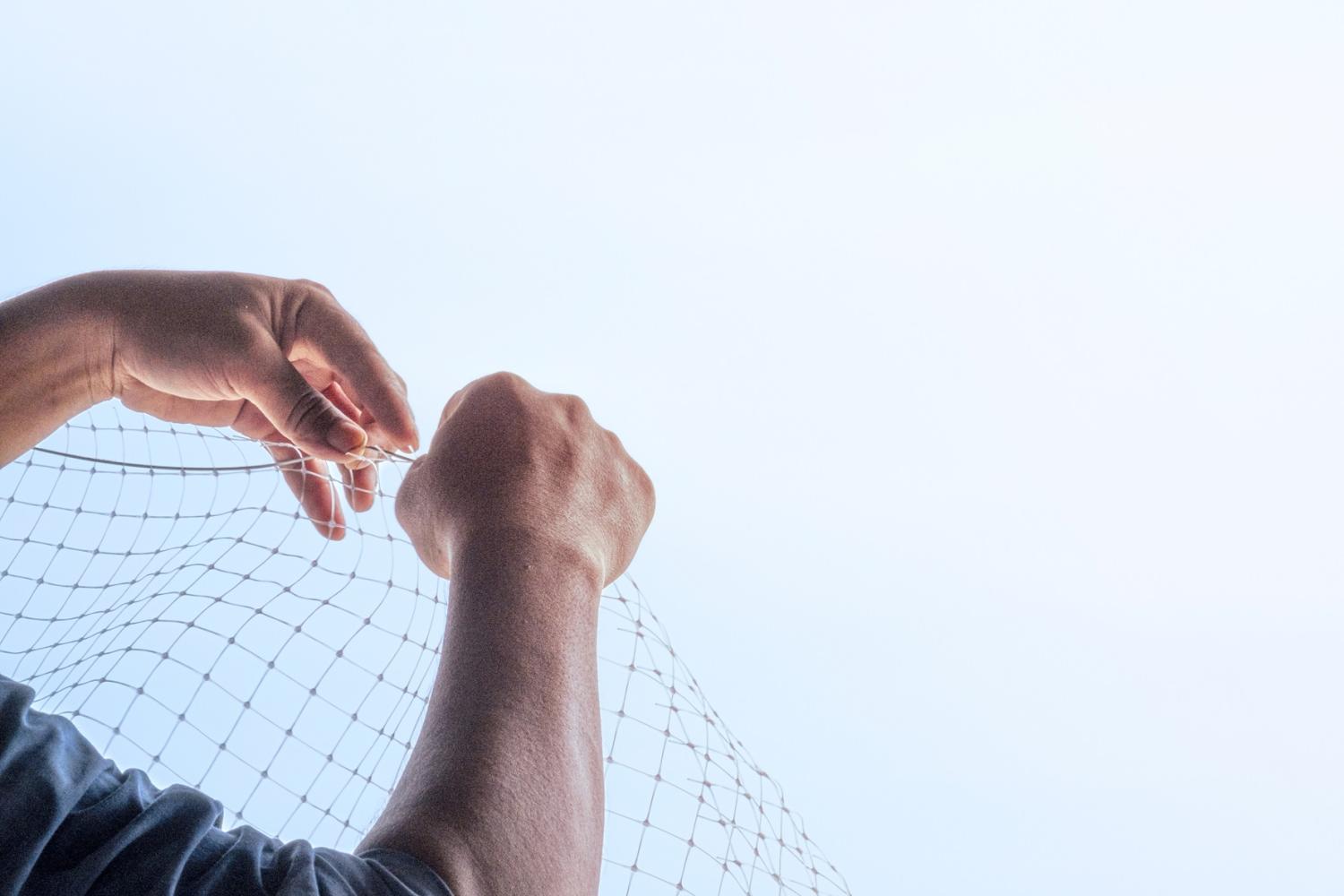 A pair of hands repairing a net, with a blue sky in the background