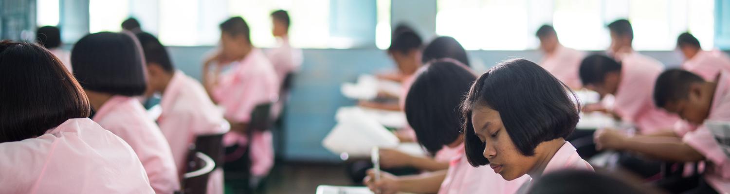 Male and female students dressed in pink uniforms sit at classroom desks taking exams.