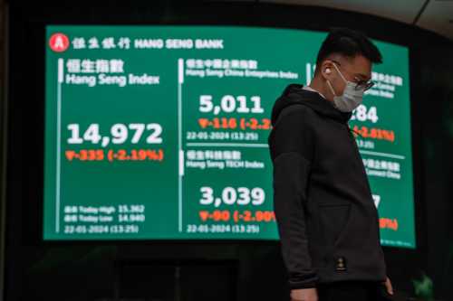 A man wears a surgical mask and looks down at the ground while standing in front of a stock display board.