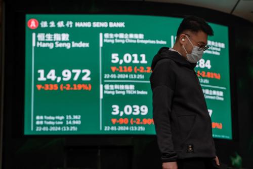 A man wears a surgical mask and looks down at the ground while standing in front of a stock display board.