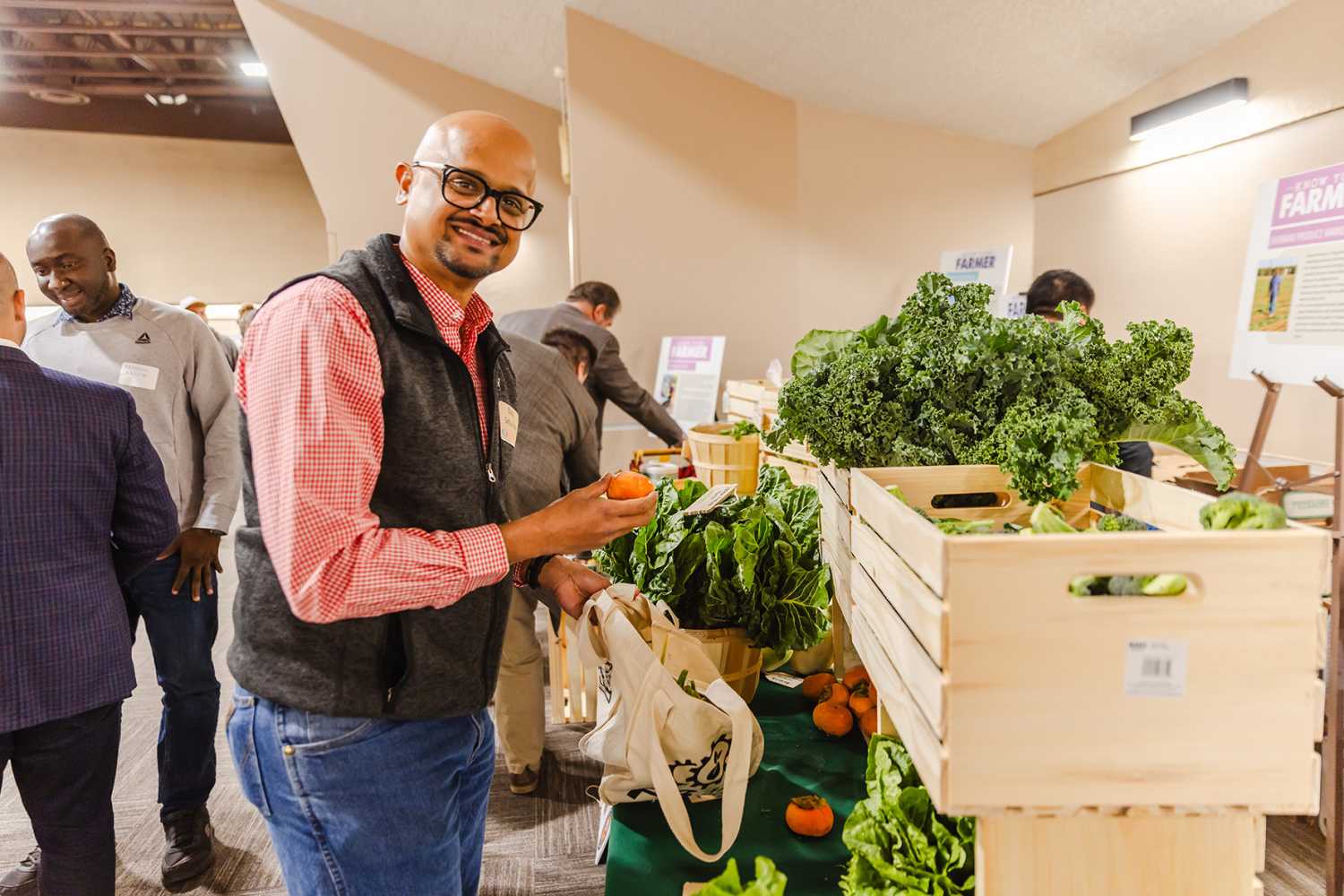 Man smiling at indoor produce table holding a vegetable