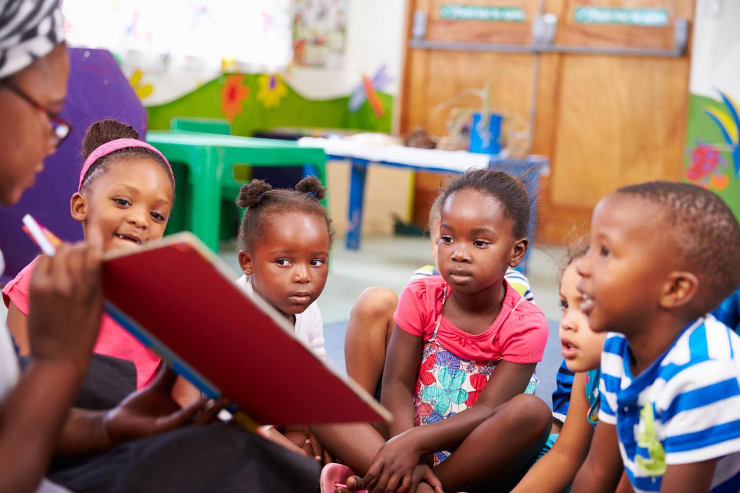 Young children sit on the floor and look at a book held by their teacher
