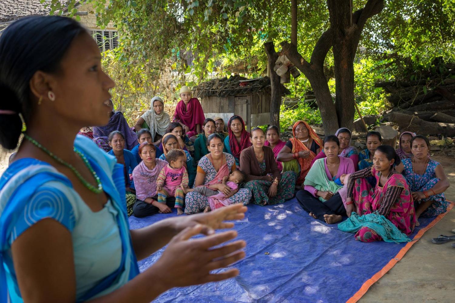 A woman gestures as she speaks to a group of women seated on the ground
