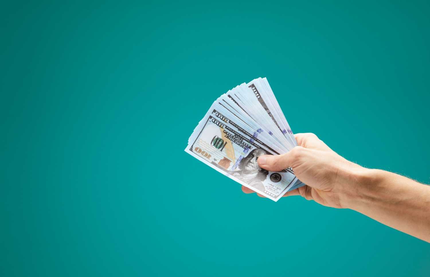 A hand holding a stack of 100 dollar bills in front of a teal background