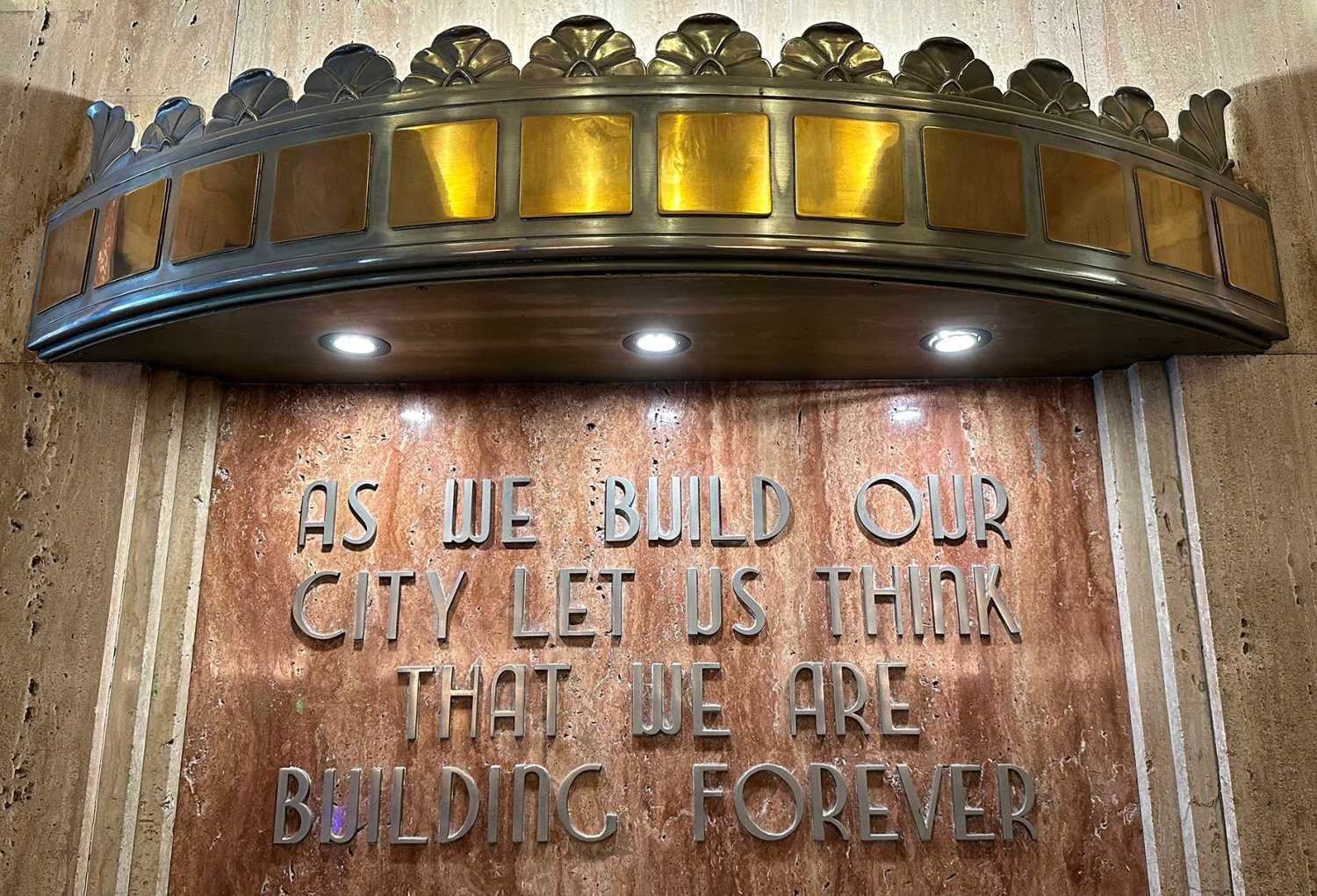Ornate brass sign that reads "As we build our city let us think that we are building forever"