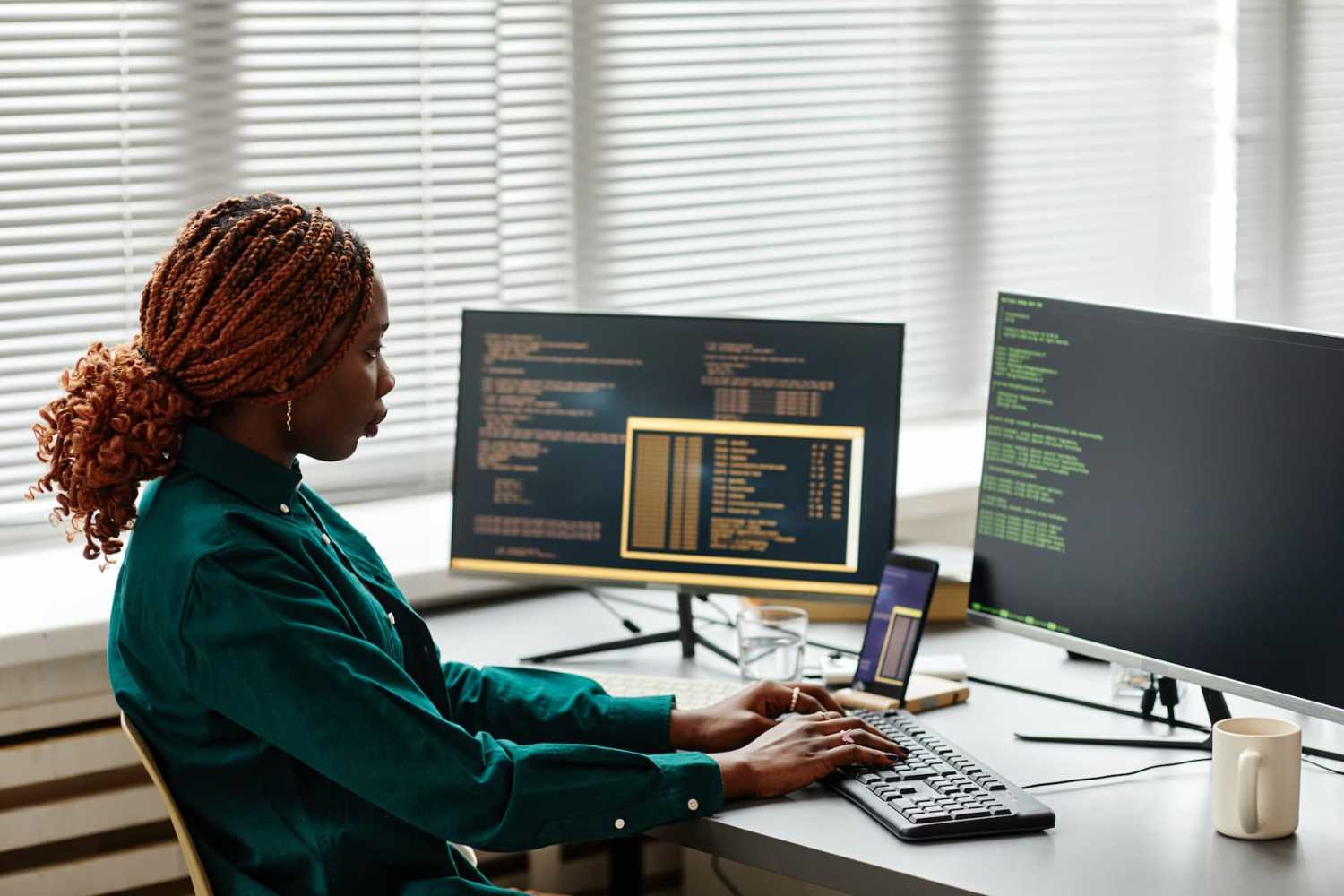 Side view of Black woman IT developer typing on keyboard with programming code on computer screen while working in office