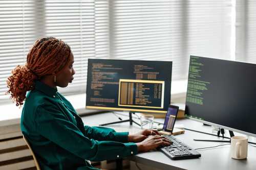 Side view of Black woman IT developer typing on keyboard with programming code on computer screen while working in office