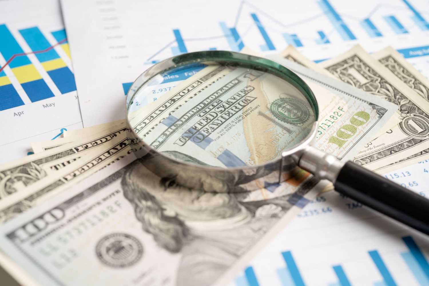 A magnifying glass lays on top of a stack of U.S. currency and printouts with financial charts
