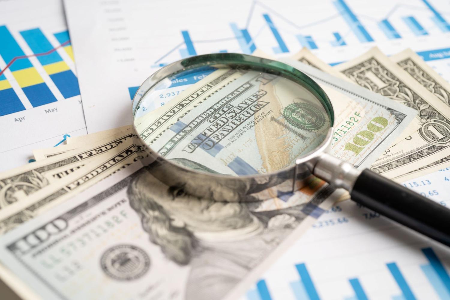 A magnifying glass lays on top of a stack of U.S. currency and printouts with financial charts