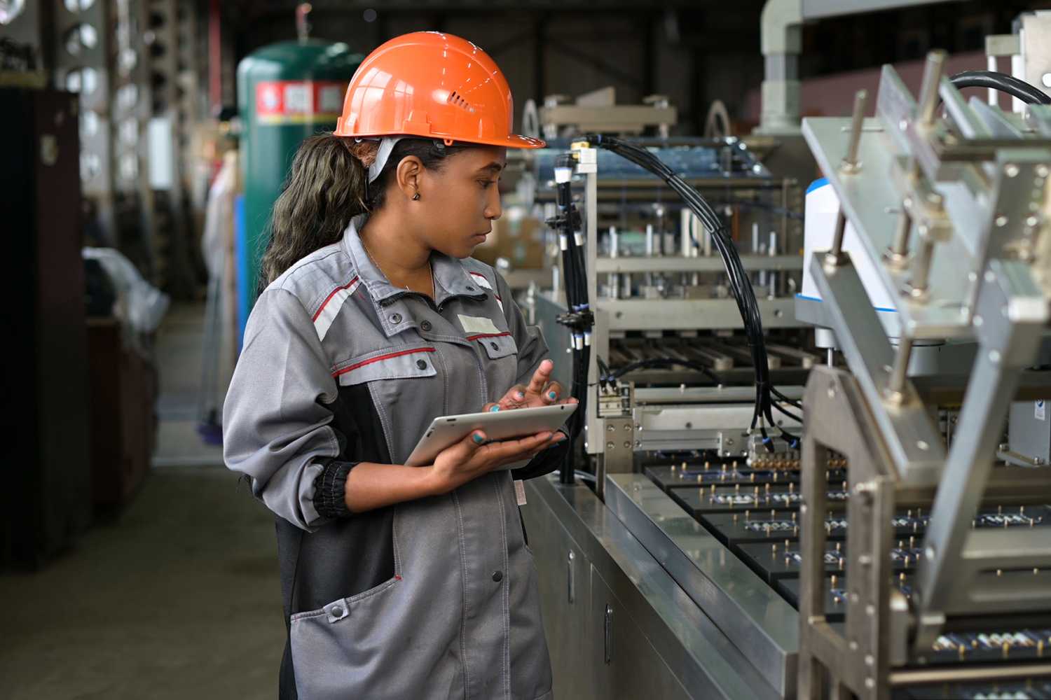A young black manufacturing worker controls an assembly line in a factory, making notes on a tablet. Young woman in hard hat watches the work process