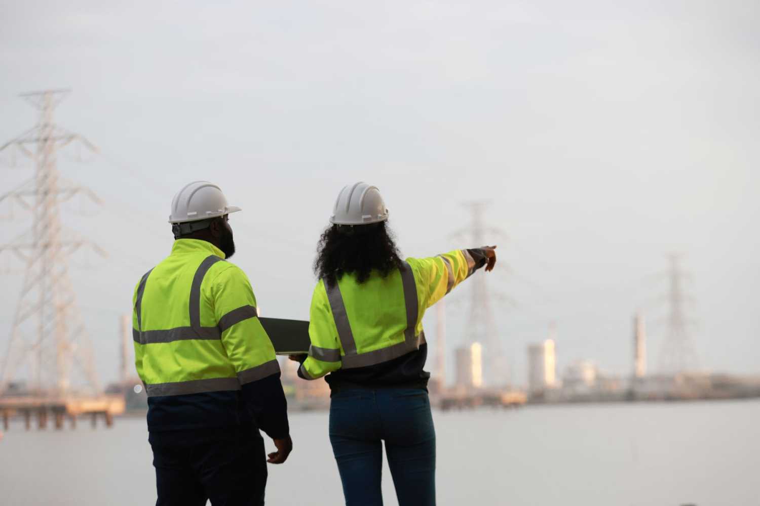 A man and woman wearing safety vests and hard hats point at powerlines across water