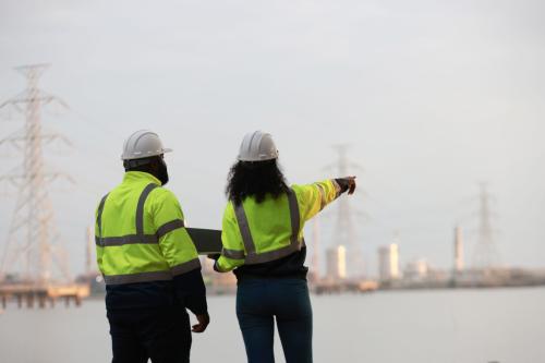 A man and woman wearing safety vests and hard hats point at powerlines across water