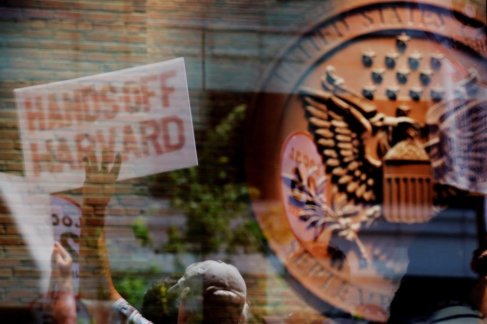Demonstrators with group Crimson Courage, who gathered to support Harvard University outside a hearing before a federal judge to order U.S. President Donald Trump's administration to restore about $2.5 billion in canceled federal grants and cease efforts to cut off research funding, are reflected in a window of the federal courthouse in Boston, Massachusetts, U.S., on July 21, 2025.