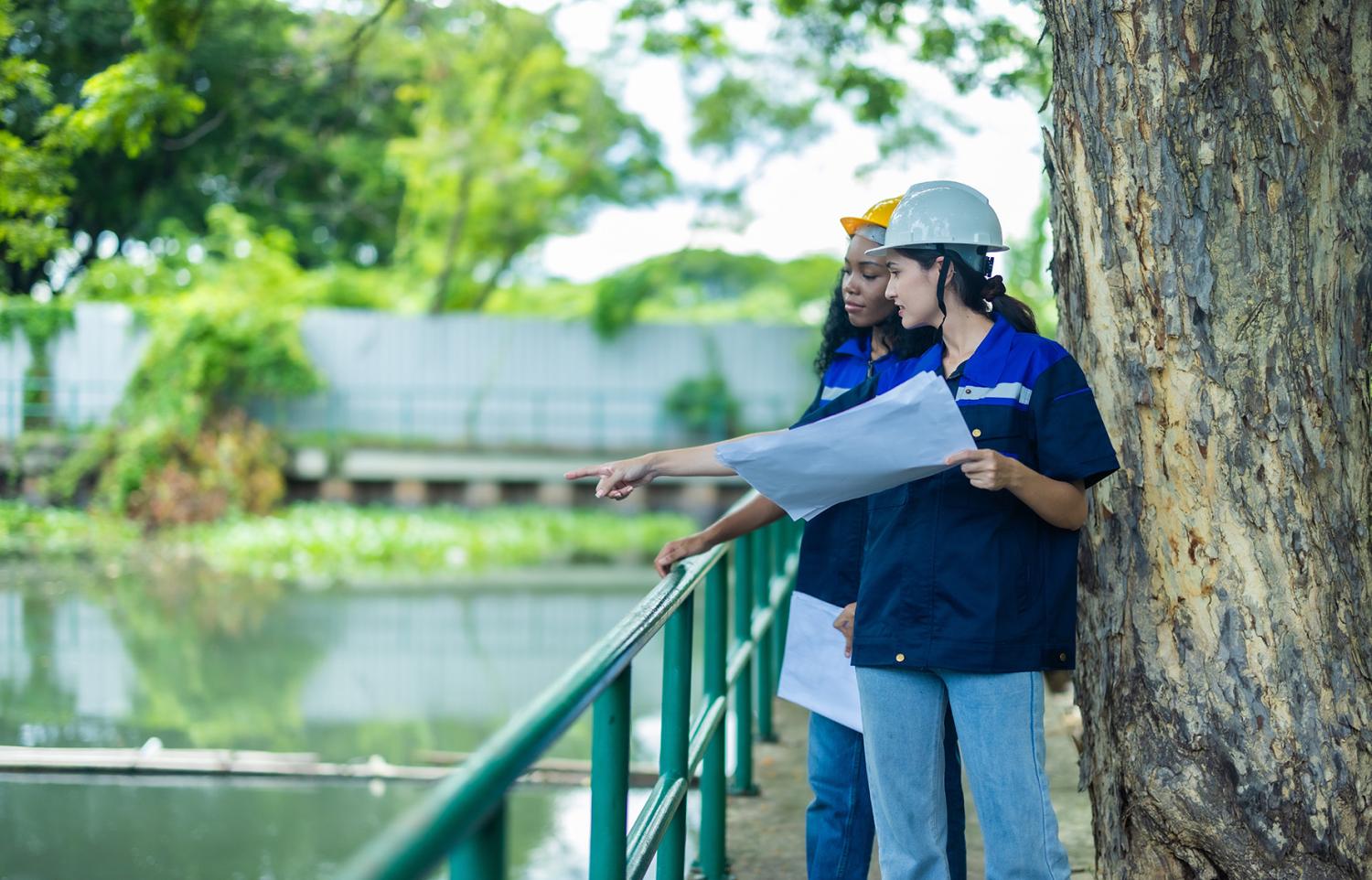 Two Environmental engineers analyze and discuss blueprints scheme. Identifying innovative strategies and sustainable practices to reduce the carbon footprint of their upcoming project.