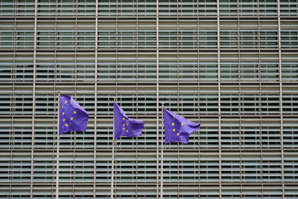 Three European Union flags, blue with a circle of yellow stars, blowing in the wind in front of a large building.