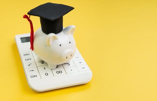 Piggy bank sitting on a calculator while wearing a graduation cap, with a yellow background.
