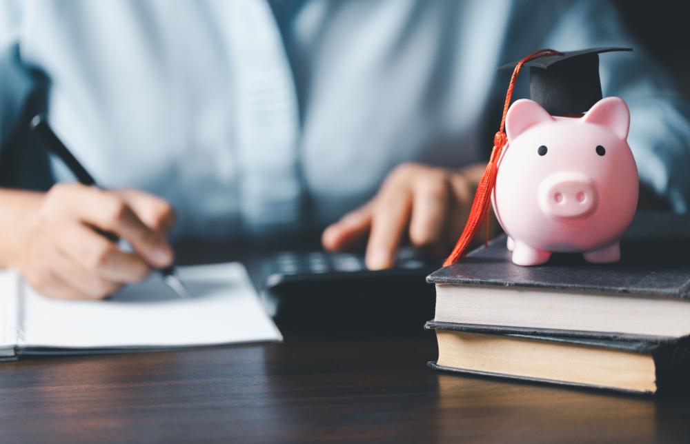 Piggy bank with a graduation cap in the foreground with a person writing and using a calculator in the background.