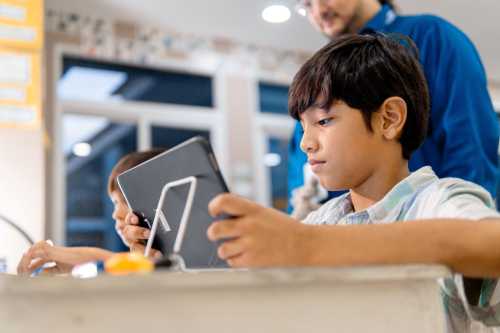 A boy sits at a desk and looks at a tablet while his teacher stands behind