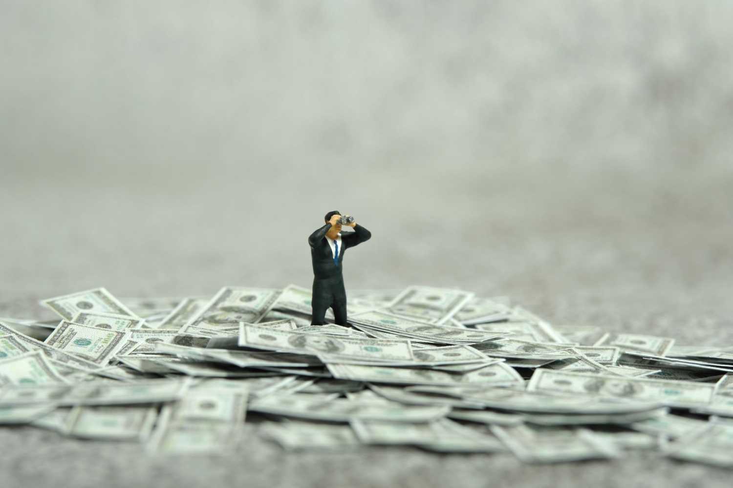 A miniature figure of a man in a suit and looking through binoculars stands in the middle of a pile of U.S. dollars