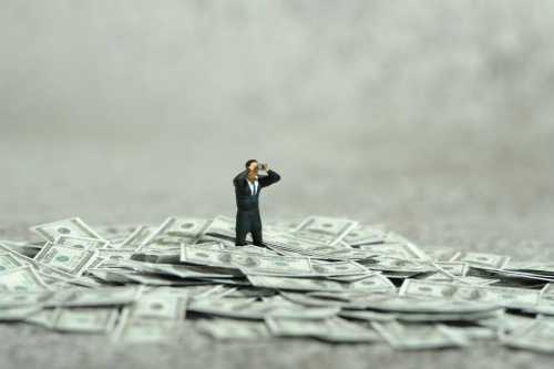 A miniature figure of a man in a suit and looking through binoculars stands in the middle of a pile of U.S. dollars
