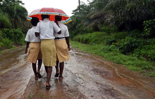 Three girls stand under a single umbrella as they walk along a muddy road