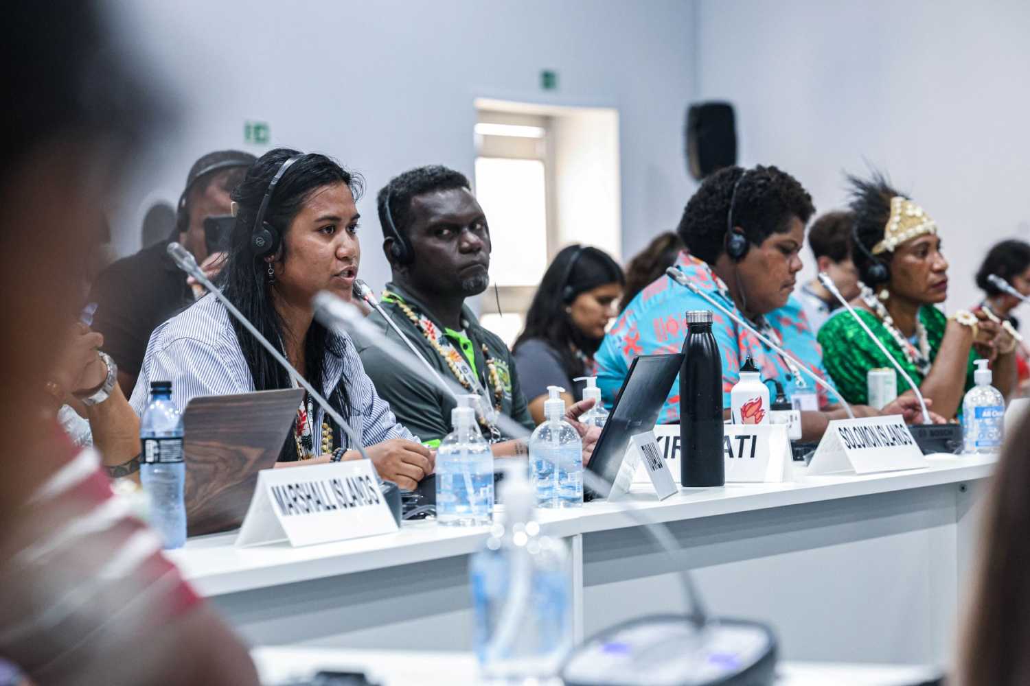 A young women sitting at a table speaks into a microphone while fellow participants look on.