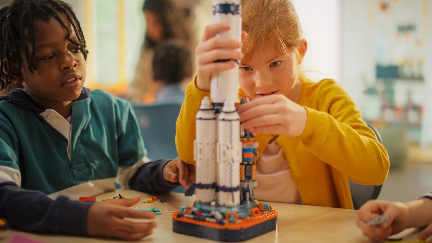 Two young students building a rocket ship model together in a classroom.