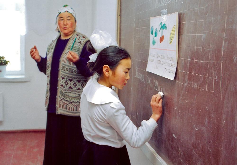 A child writes on a blackboard while her teacher watches.