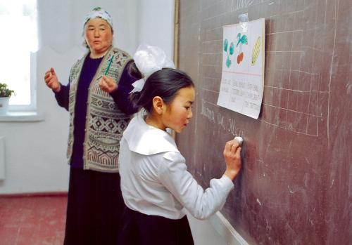 A child writes on a blackboard while her teacher watches.