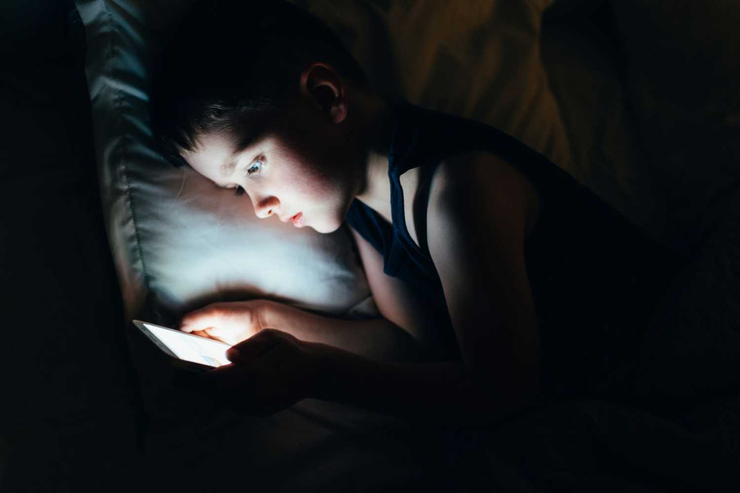 A young boy looks at a glowing phone screen while lying in bed