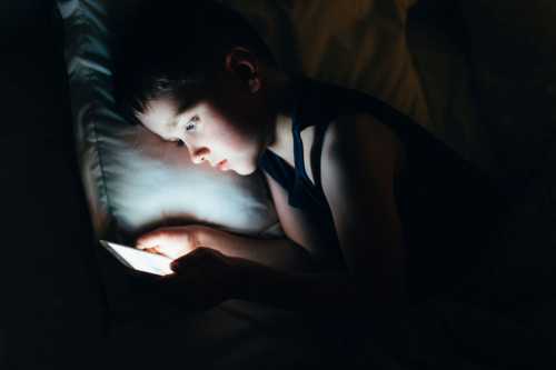 A young boy looks at a glowing phone screen while lying in bed