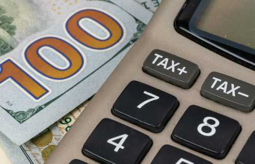 A calculator with plus and minus tax buttons on top of $100 dollar bill.