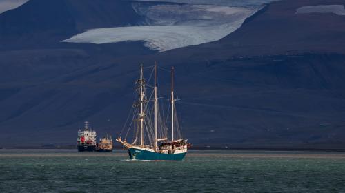 Two sailing vessels navigate the tranquil Arctic waters of Svalbard, with majestic glaciers visible in the background under a bright sky.