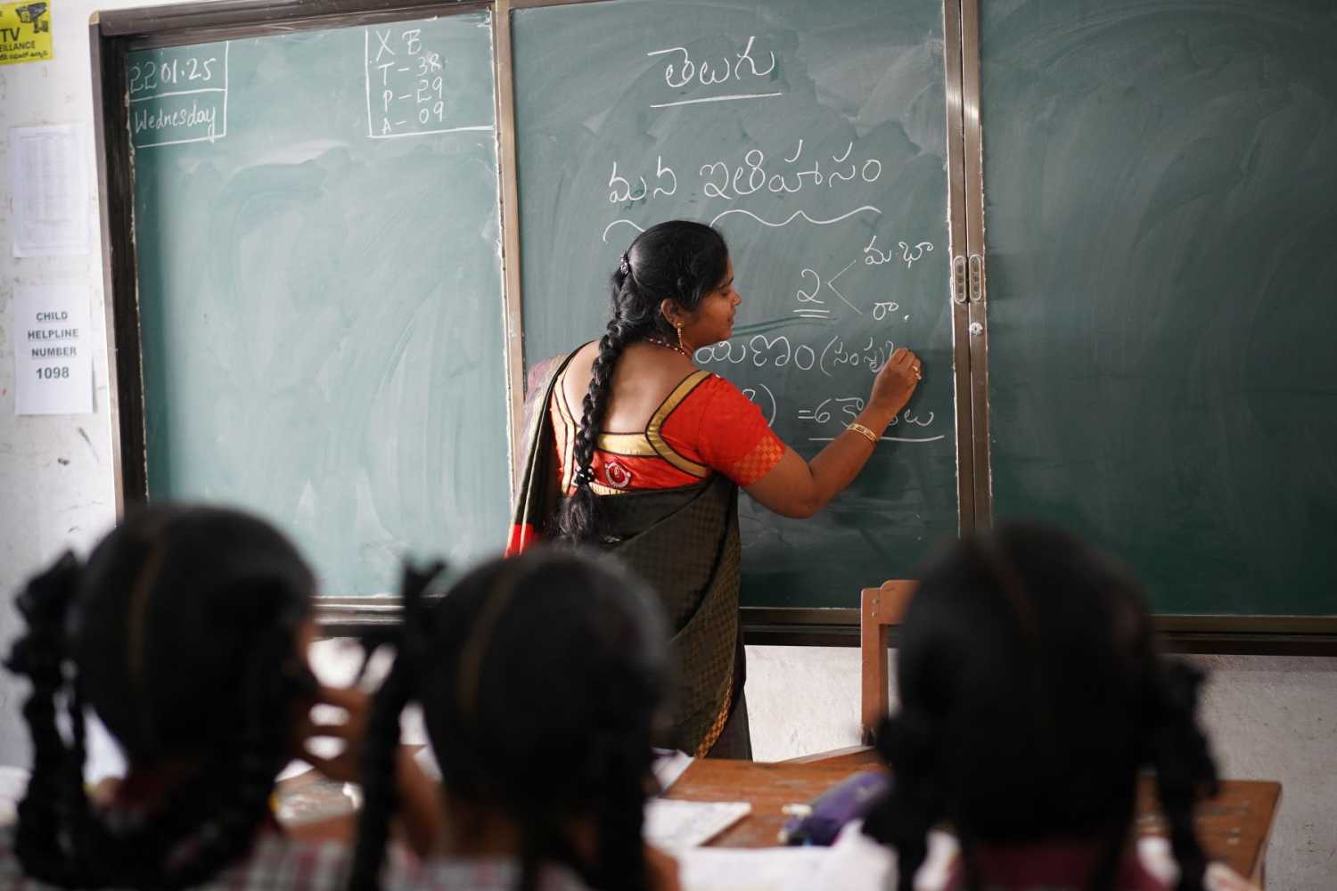 A teacher writes on a chalkboard at the front of a classroom.