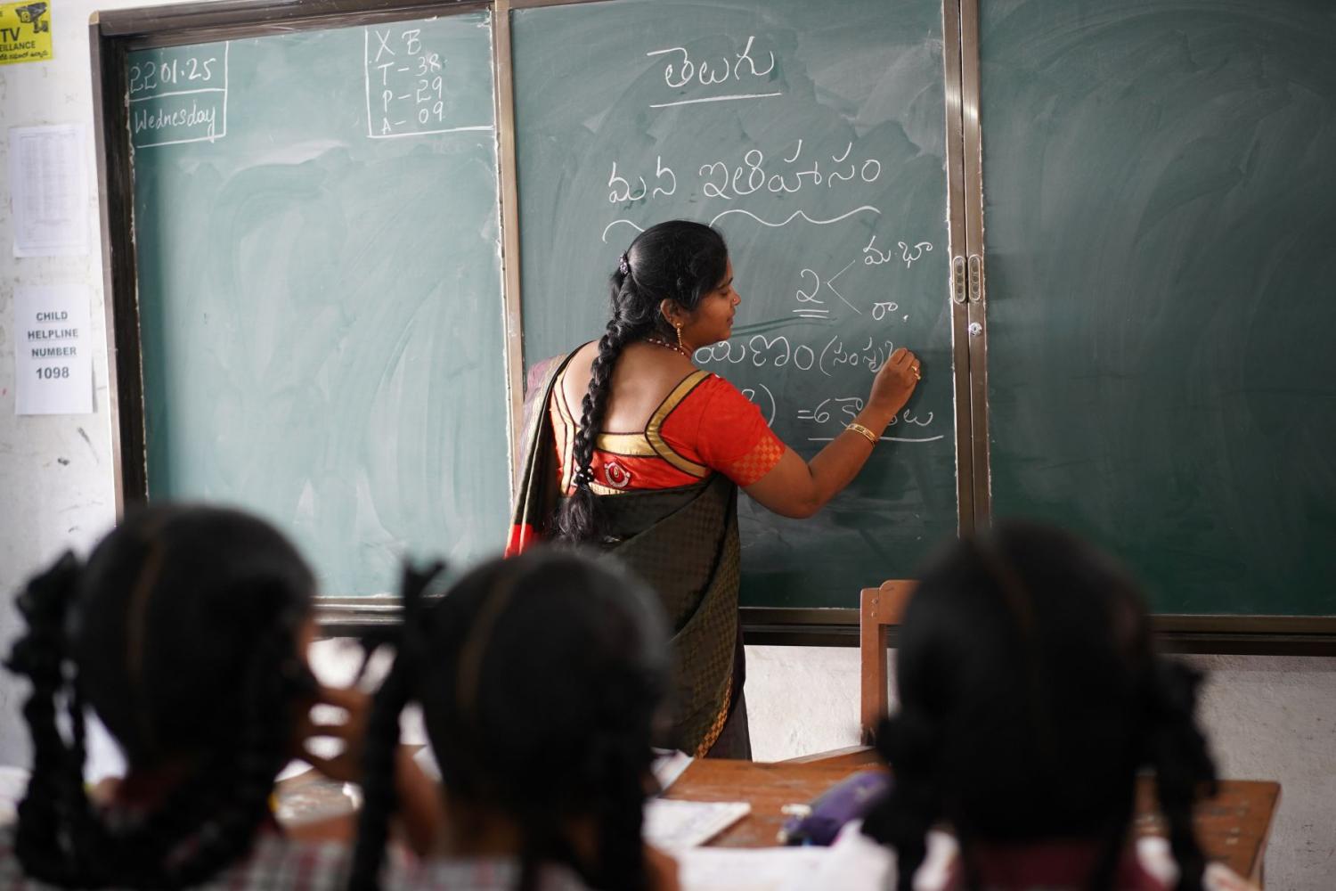 A teacher writes on a chalkboard at the front of a classroom.