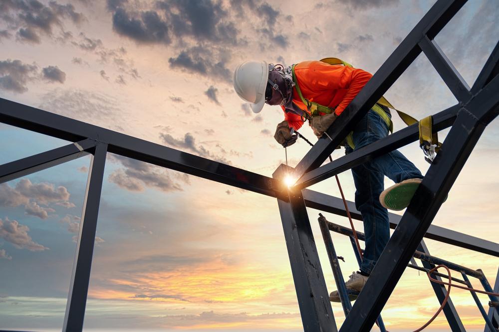 A welder is welding steel on a steel roof truss. Working at height equipment. Fall arrestor device for worker with hooks for safety body harness. Worker in construction site.