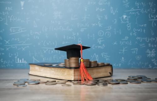 Coins and a graduation cap on top of a book, in front of a chalkboard.