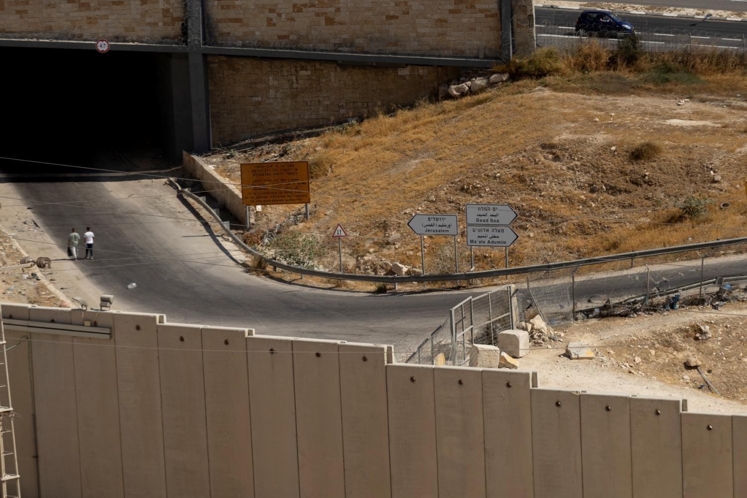 People walk by signs leading to Jerusalem and an Israeli settlement in the West Bank called Ma'ale Adomim, which the Israeli government announced it would expand on August 22, 2025.