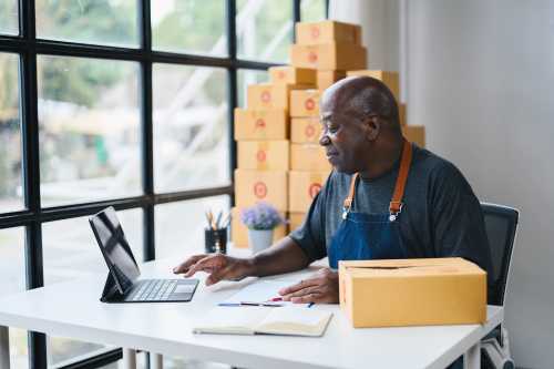 Black man working on a tablet in a home office surrounded by cardboard boxes, representing a small business owner or e-commerce entrepreneur preparing for shipping.