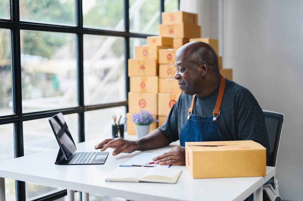 Black man working on a tablet in a home office surrounded by cardboard boxes, representing a small business owner or e-commerce entrepreneur preparing for shipping.
